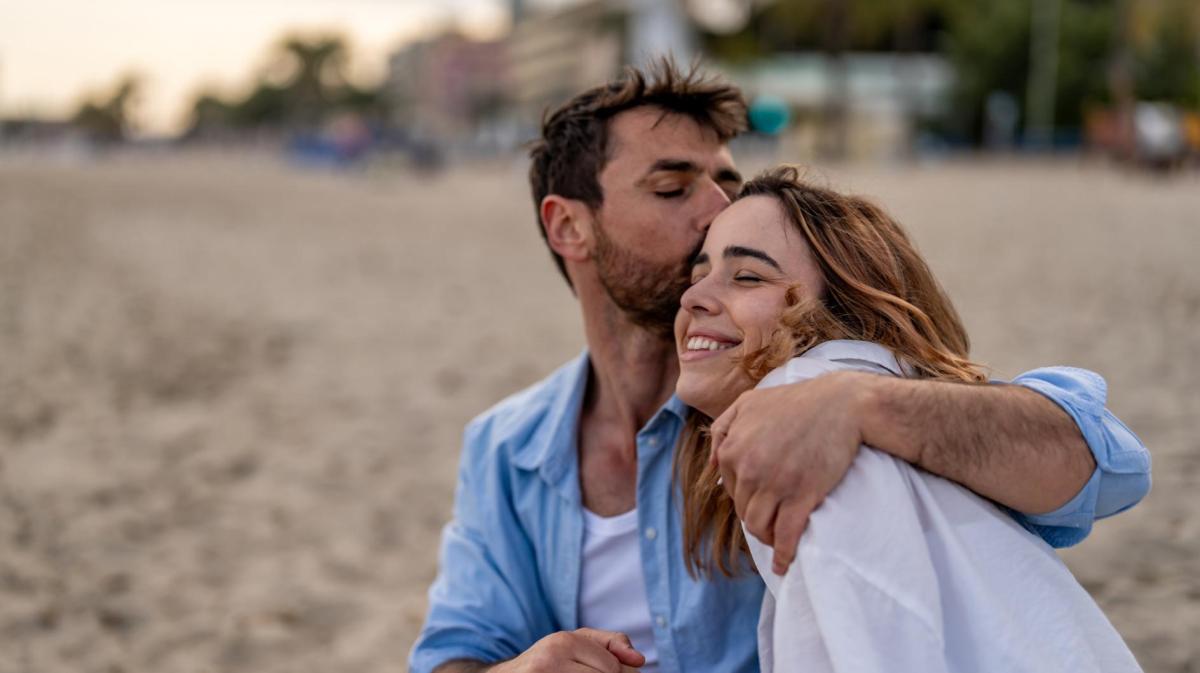 Ein Mann mit Dreitagebart in offenem Jeanshemd küsst eine lachende Frau mit welligen, braunen Haaren auf die Stirn am Sandstrand.