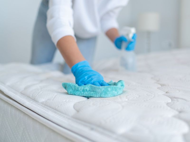 Frau säubert eine Matratze mit einem Tuch und einer Sprühflasche.