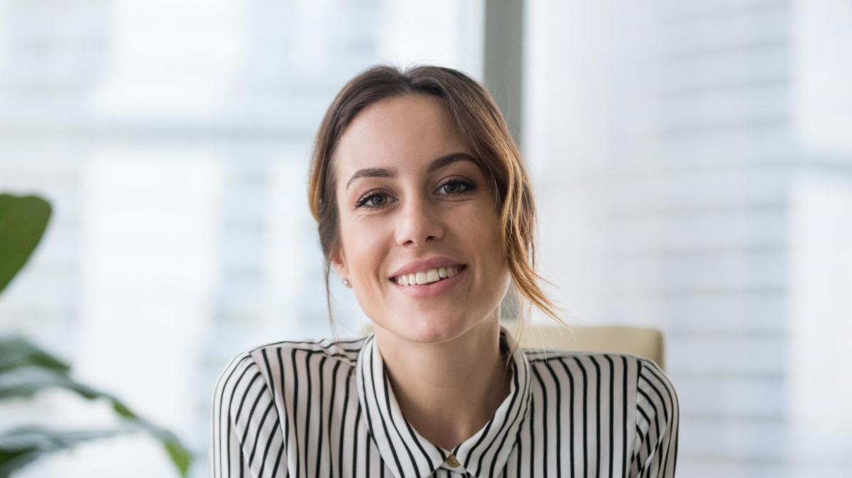 Junge Frau mit zusammengesteckten Haaren und einem gestreiften Hemd lächelt am Schreibtisch.