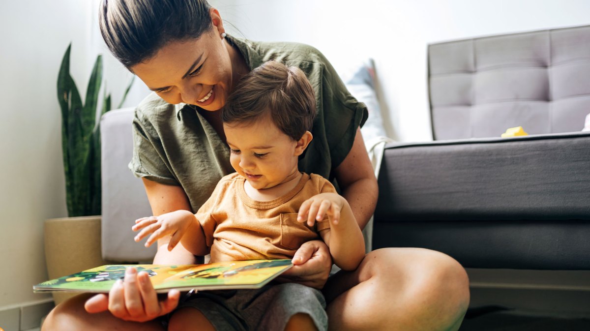 Mutter und Sohn sitzen auf dem Fußboden vor einer Couch, der Sohn auf dem Schoß der Mutter, und beide schauen gemeinsam in ein Buch.
