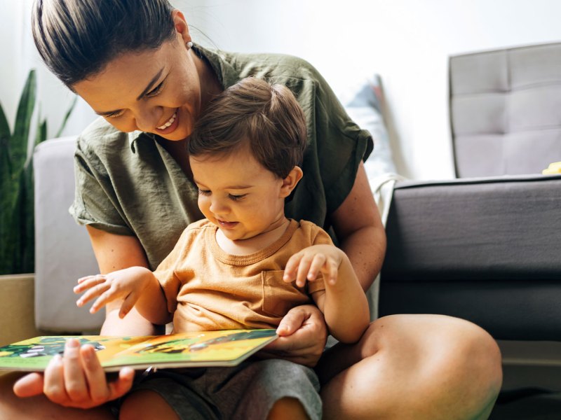 Mutter und Sohn sitzen auf dem Fußboden vor einer Couch, der Sohn auf dem Schoß der Mutter, und beide schauen gemeinsam in ein Buch.