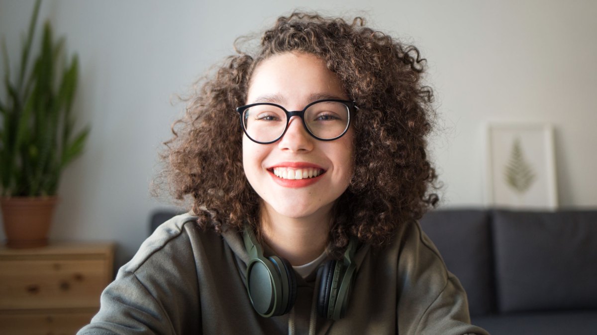 Teenagerin sitzt mit Köpfhörern um den Hals in ihrem Zimmer und lacht glücklich in die Kamera.