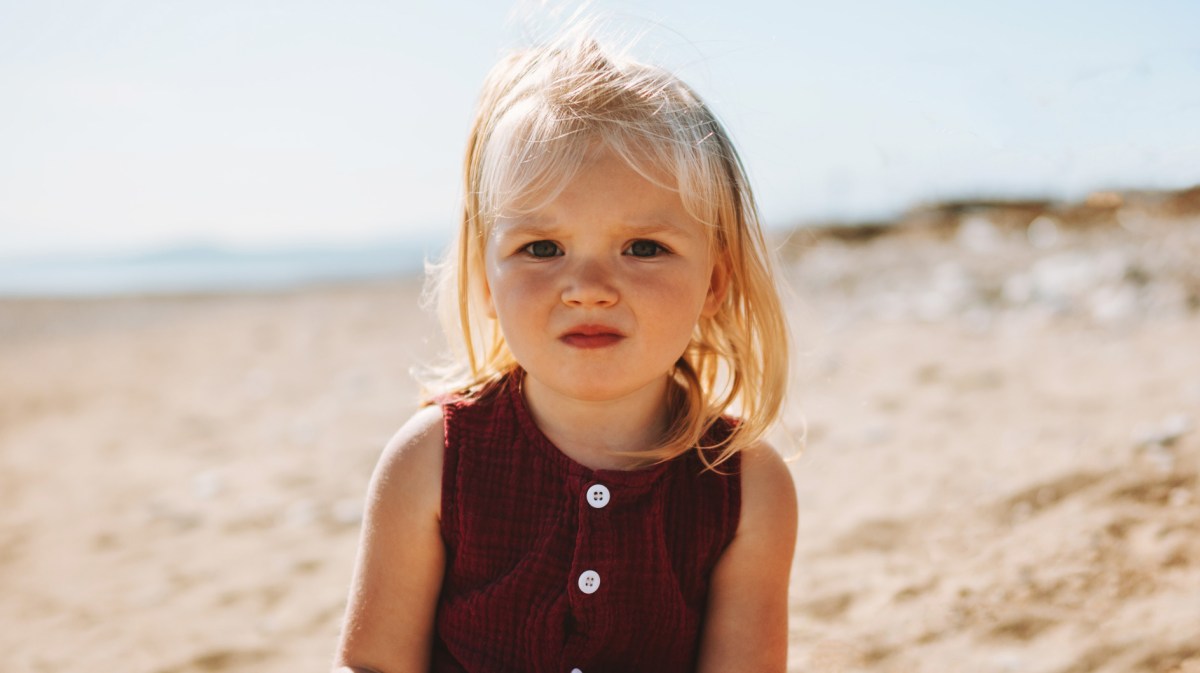 Porträt eines Mädchens, 2 Jahre alt, das in der Sonne am Strand sitzt und ernst schaut.