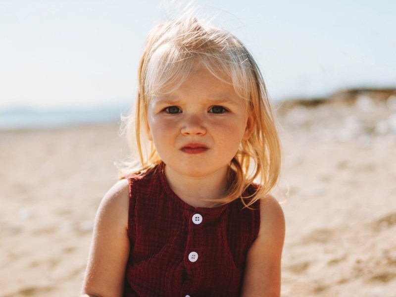 Porträt eines Mädchens, 2 Jahre alt, das in der Sonne am Strand sitzt und ernst schaut.