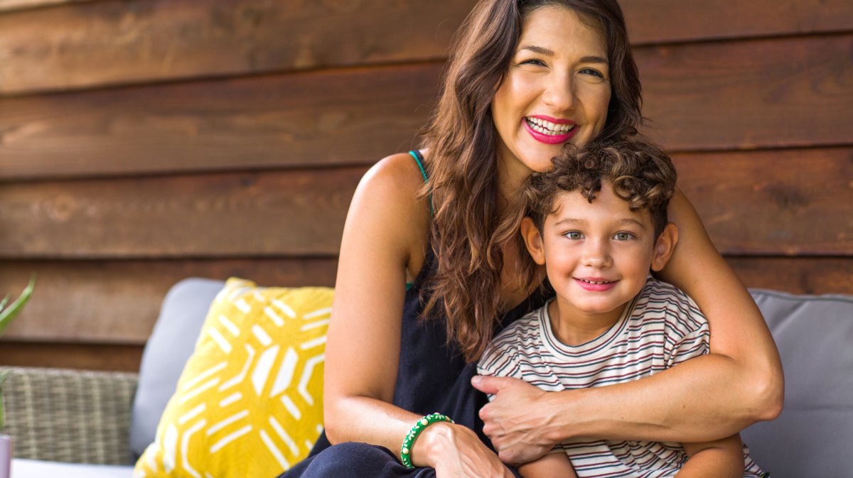 Mutter umarmt Sohn auf der Terrasse vor einem Holzhaus.