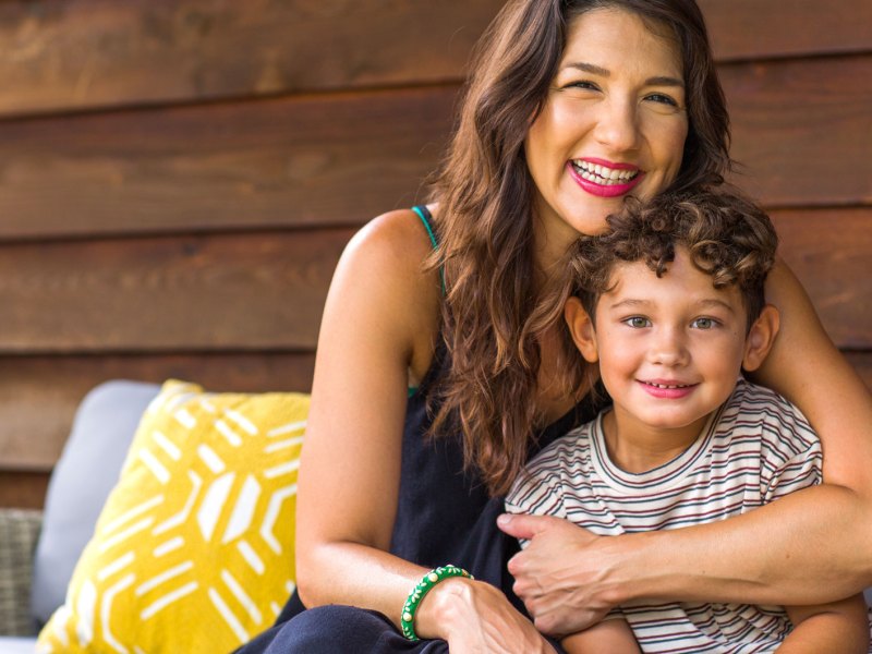 Mutter umarmt Sohn auf der Terrasse vor einem Holzhaus.