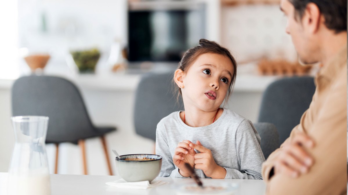 Mädchen sitzt mit seinem Vater am Frühstückstisch und bettelt um etwas.