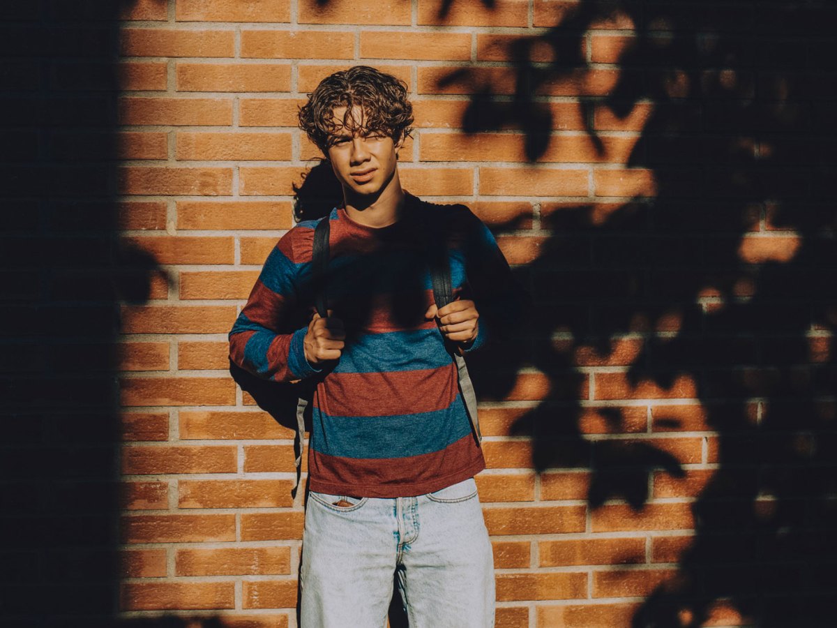 Junge im Teenageralter steht vor einer gemauerten Wand im Halbschatten und schaut ernst in Richtung Kamera. Er trägt Jeans, Sweatshirt und Rucksack.