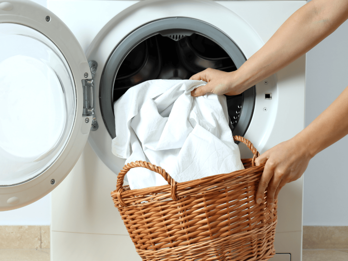Person gibt weiße Wäsche aus einem Strohkorb in die Waschmaschine