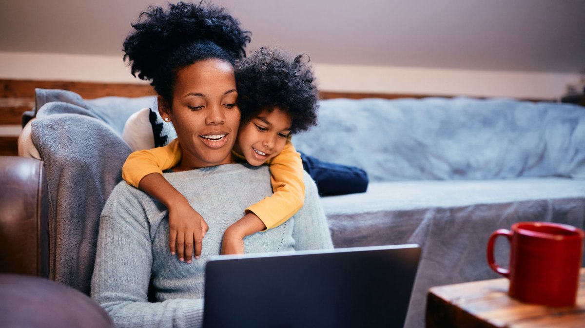 Mutter und Tochter sitzen im Wohnzimmer und schauen etwas am Laptop. Die Mutter sitzt auf dem Fußboden, an die Couch gelehnt, die Tochter schlägt von hinten ihre Arme um die Mutter und schaut ihr über die Schulter.