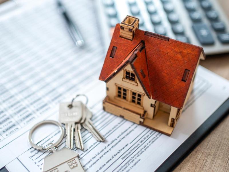 Miniaturhaus mit rotem Dach steht auf Vertragsdokumenten neben Hausschlüsseln, Kugelschreiber und Taschenrechner.