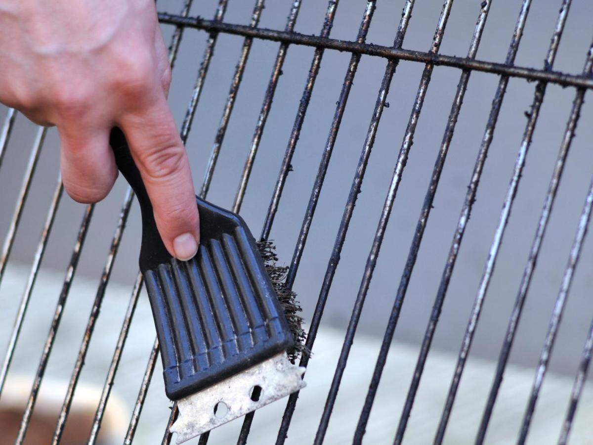 Eine Hand mit Grillb&uuml;rste reinigt einen stark verschmutzten Grillrost &uuml;ber hellem Boden im Freien bei Tageslicht.