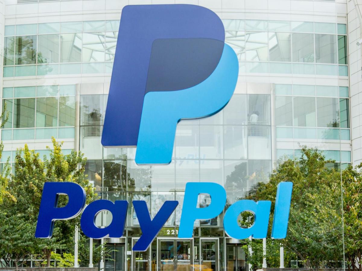 Das große PayPal-Logo in Blau vor dem modernen, verglasten Firmensitz mit weißen Fassaden und grünen Bäumen im Eingangsbereich.