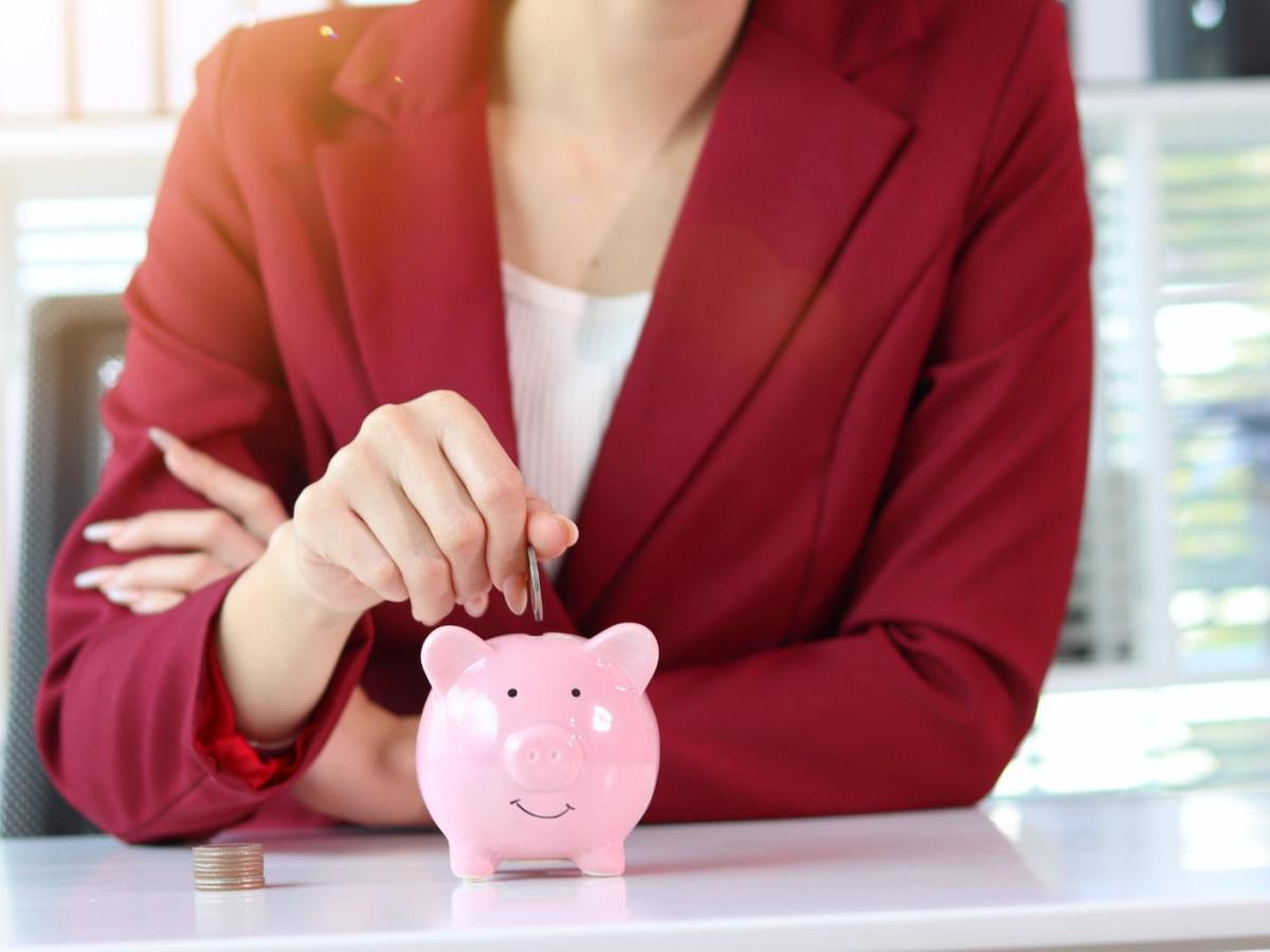 Eine l&auml;chelnde Frau im bordeauxroten Blazer steckt eine M&uuml;nze in ein rosa Sparschwein auf einem wei&szlig;en Schreibtisch.