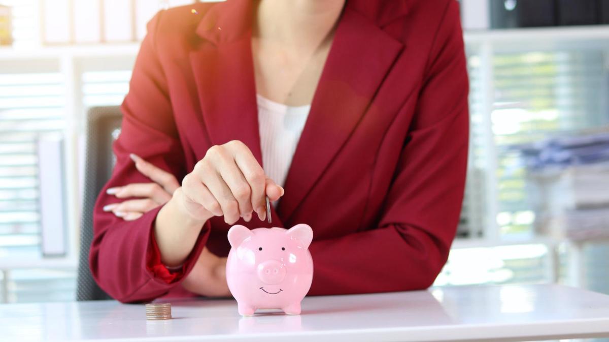 Eine lächelnde Frau im bordeauxroten Blazer steckt eine Münze in ein rosa Sparschwein auf einem weißen Schreibtisch.