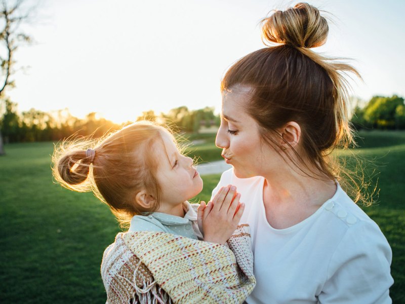 Mutter und Tochter (4) draußen im Park. Die Mutter trägt die Tochter, die in eine Decke gewickelt ist.