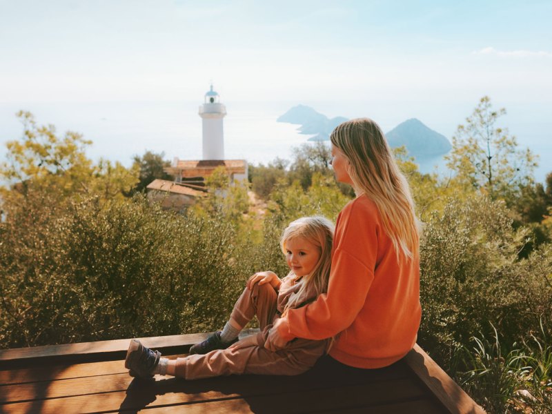 Mutter und Kind auf Reisen, sitzen auf einem Felsvorsprung, im Hintergrund erkennt man einen Turm und Berge im Meer.