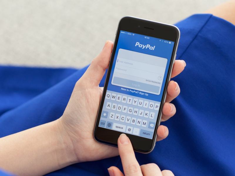 Frau mit blauem Kleid tippt mit ihrem Finger auf einem Handy, auf dem die Paypal App geöffnet ist.