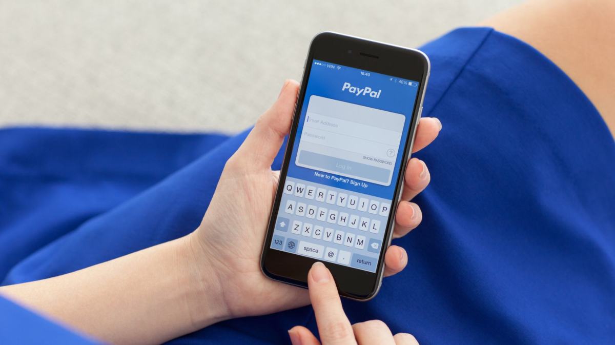 Frau mit blauem Kleid tippt mit ihrem Finger auf einem Handy, auf dem die Paypal App geöffnet ist.