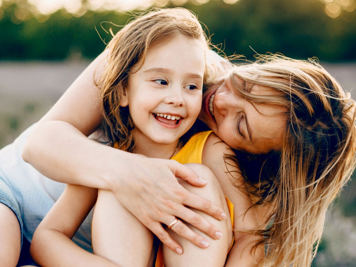 Mutter und Tochter sitzen auf einer Wiese an einem Sommerabend. Die Mutter umarmt ihr Kind von hinten, das lacht glücklich.
