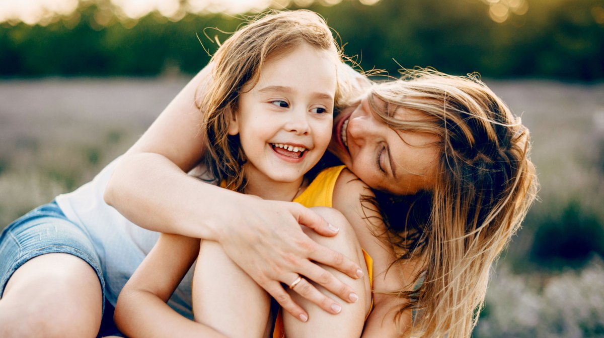 Mutter und Tochter sitzen auf einer Wiese an einem Sommerabend. Die Mutter umarmt ihr Kind von hinten, das lacht glücklich.