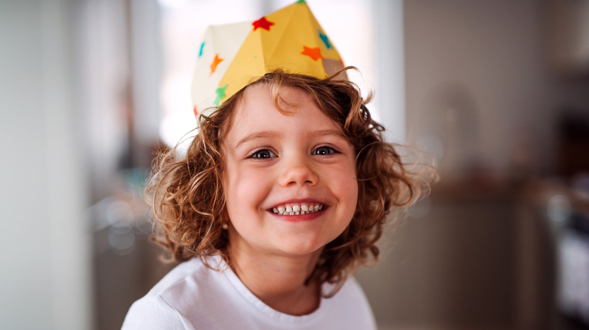 Kleines Kind, ca. 5 Jahre alt, strahlt in die Kamera und trägt eine Krone aus Papier.