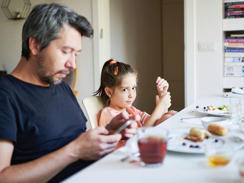 Vater und Tochter sitzen am Frühstückstisch. Der Vater schaut währenddessen auf sein Smartphone.