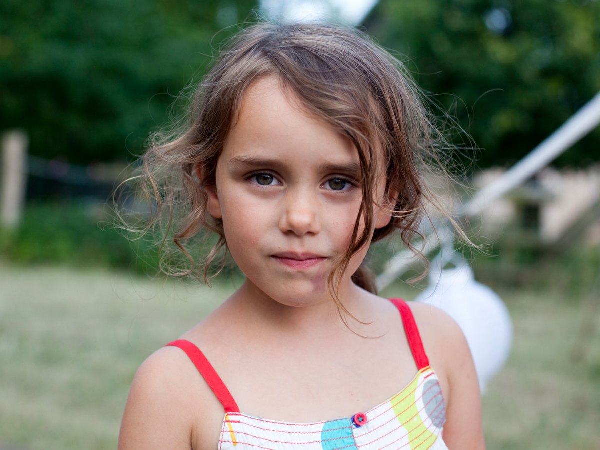 Portr&auml;t eines jungen M&auml;dchens, das ernst in die Kamera schaut, w&auml;hrend es vor einem Spielplatz steht.