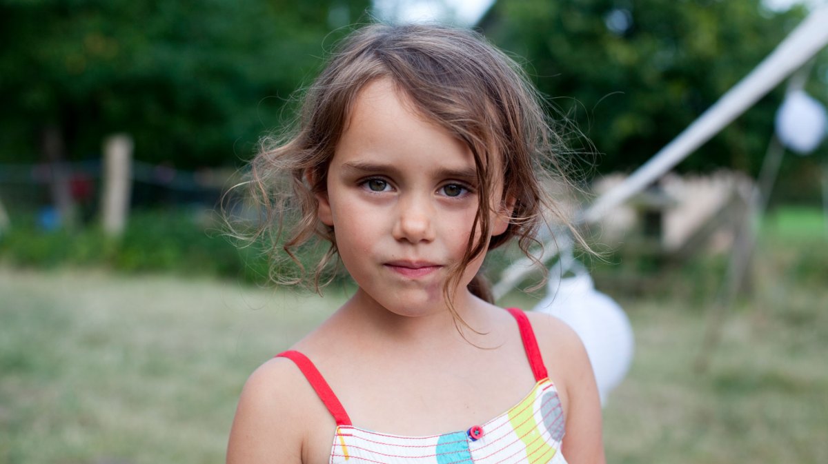 Porträt eines jungen Mädchens, das ernst in die Kamera schaut, während es vor einem Spielplatz steht.