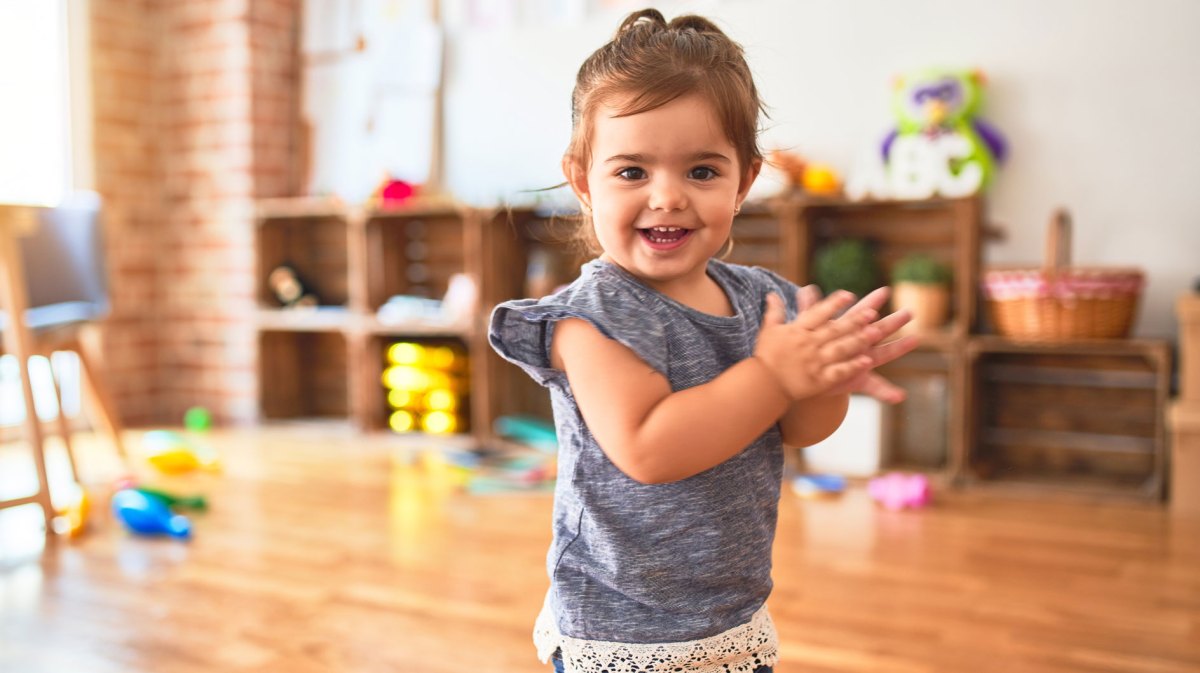 Süßes kleines Mädchen steht in einem großen Raum voller Spielsachen, lacht in die Kamera und klatscht.