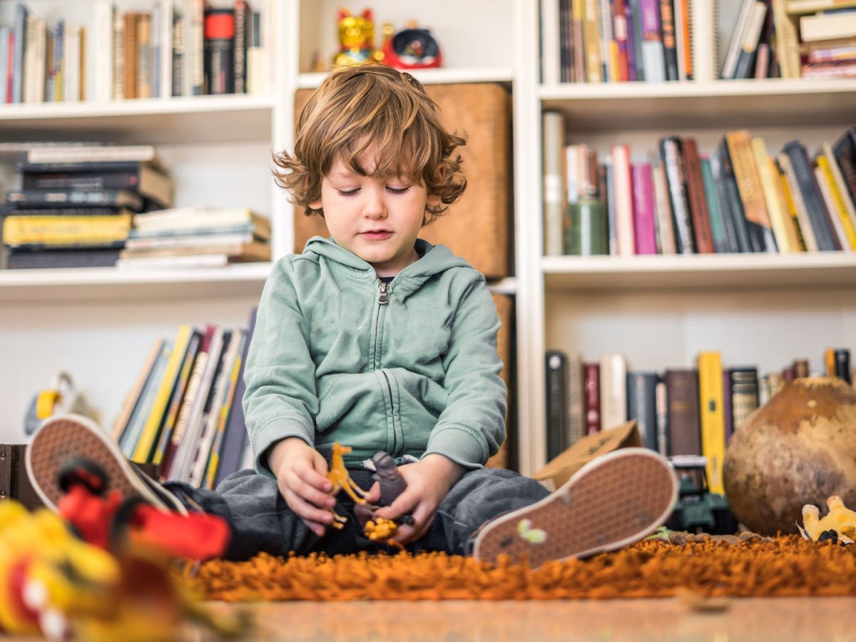 Kleiner junge sitzt auf dem Teppich umgeben von Spielsachen und schaut unglücklich auf das in seinen Händen.