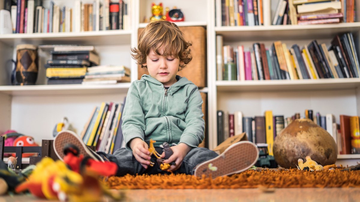 Kleiner junge sitzt auf dem Teppich umgeben von Spielsachen und schaut unglücklich auf das in seinen Händen.