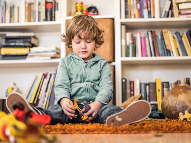 Kleiner junge sitzt auf dem Teppich umgeben von Spielsachen und schaut unglücklich auf das in seinen Händen.
