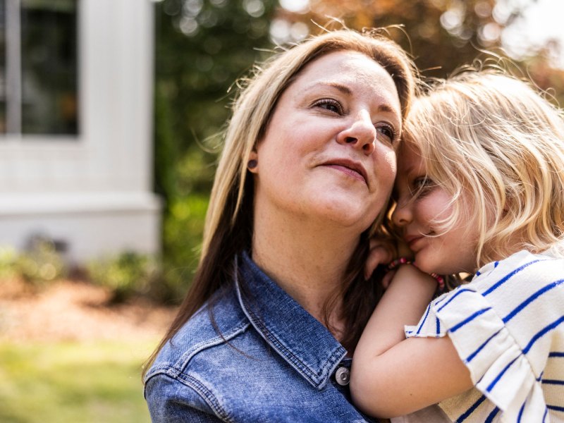 Ältere Mutter trägt Tochter, ca. 4 Jahre alt, auf dem Arm. Sie stehen draußen, im Hintergrund ist ein Einfamilienhaus angedeutet.