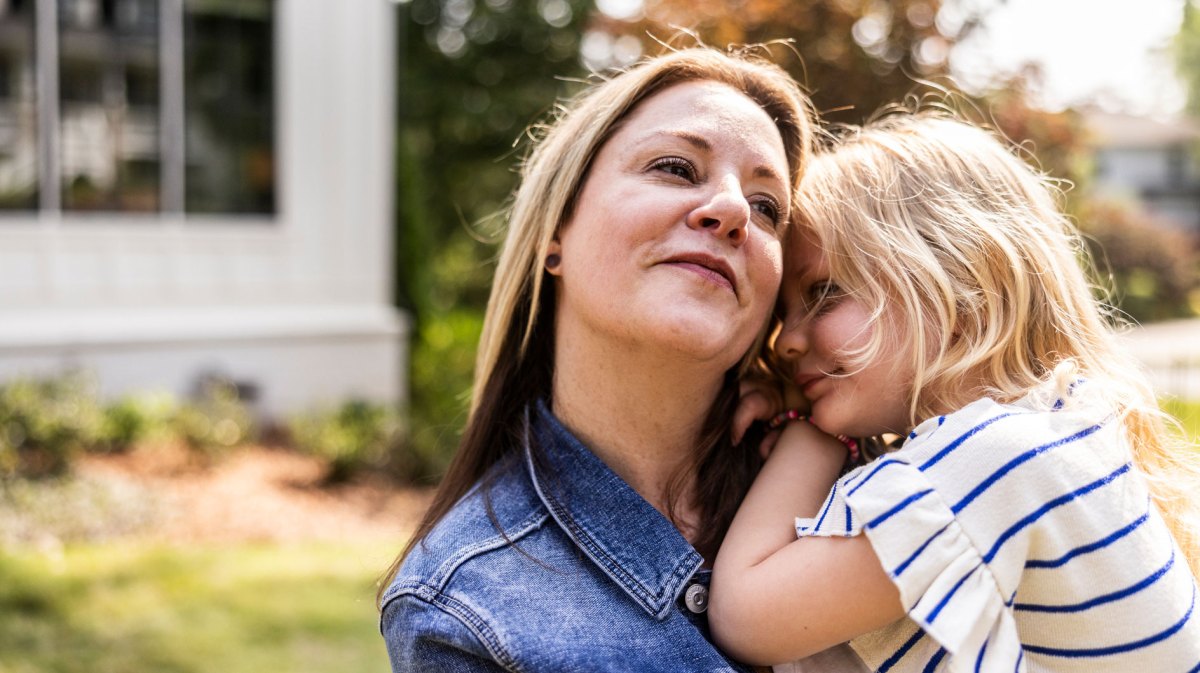 Ältere Mutter trägt Tochter, ca. 4 Jahre alt, auf dem Arm. Sie stehen draußen, im Hintergrund ist ein Einfamilienhaus angedeutet.