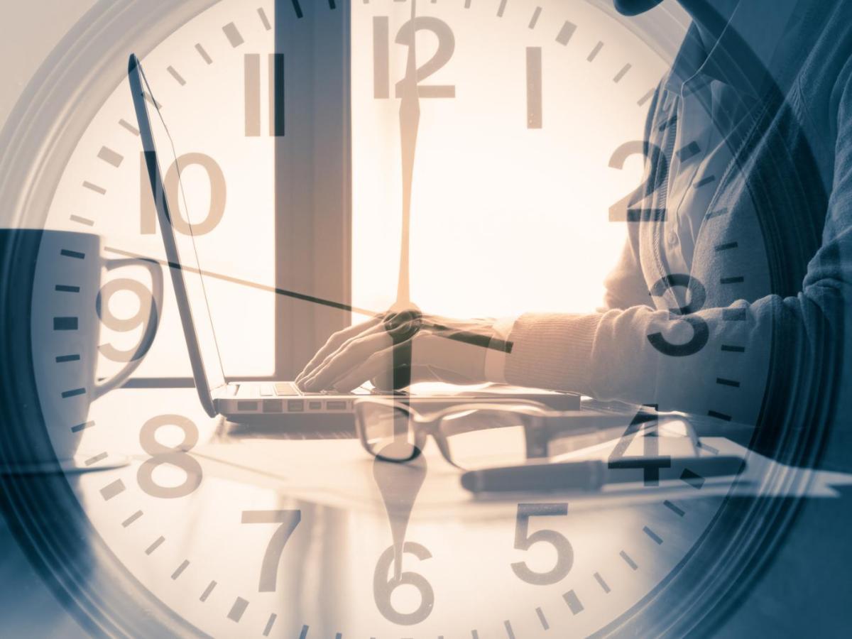 Schattenhafte Frau tippt konzentriert auf Laptop, w&auml;hrend gro&szlig;e transparente Uhr &uuml;ber dem Bild die Zeit symbolisiert.