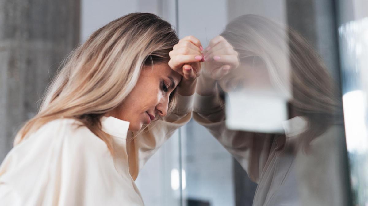 Blonde Frau lehnt sich auf der Arbeit gegen eine Glaswand und stützt dabei ihren Kopf auf ihrem Arm ab.