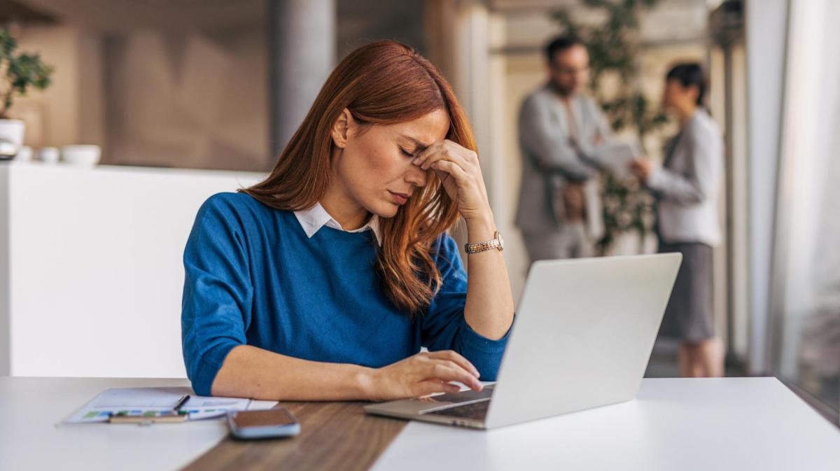 Frau fasst sich an die Stirn an ihrem Schreibtisch im Büro. Im Hintergrund stehen Kolleginnen und Kollegen.