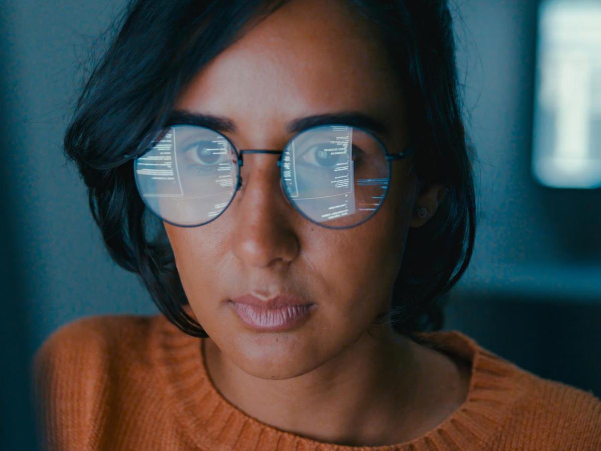 Frau mit dunklen Haaren und Brille sitzt nah vor einem Bildschirm und arbeitet lange.