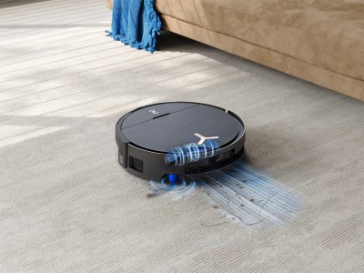 Die ZeroTangle 3.0-Technologie verhindert, dass sich Haare in den Bürsten verfangen – für eine gleichbleibend starke Reinigungsleistung und weniger Wartungsaufwand. Schwarzer Saugroboter mit glänzender Oberfläche entfernt auf einem hellgrauen Teppich unter Sonnenlicht effektiv Staub und Haare.