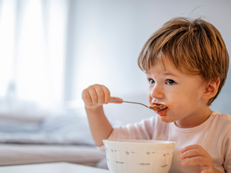 Kleinkind, etwa 2 Jahre, sitzt am Esstisch und isst mit einem Löffel aus einer Schüssel.