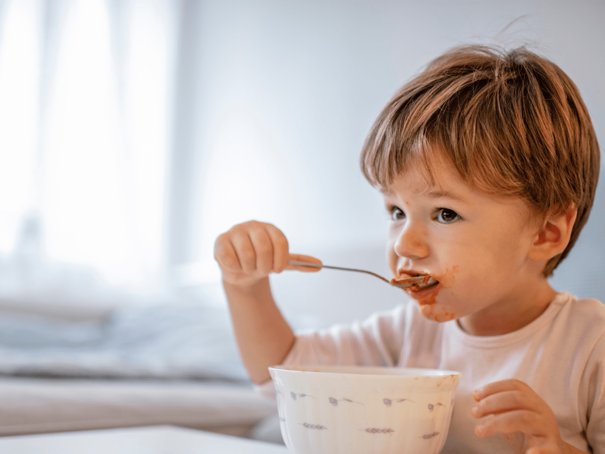 Kleinkind, etwa 2 Jahre, sitzt am Esstisch und isst mit einem Löffel aus einer Schüssel.