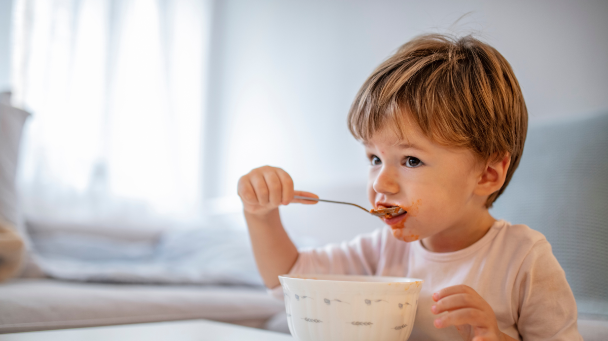 Kleinkind, etwa 2 Jahre, sitzt am Esstisch und isst mit einem Löffel aus einer Schüssel.