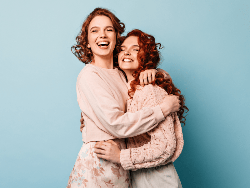 2 junge Frauen stehen vor einem hellblauen Hintergrund, freuen sich und nehmen sich in den Arm