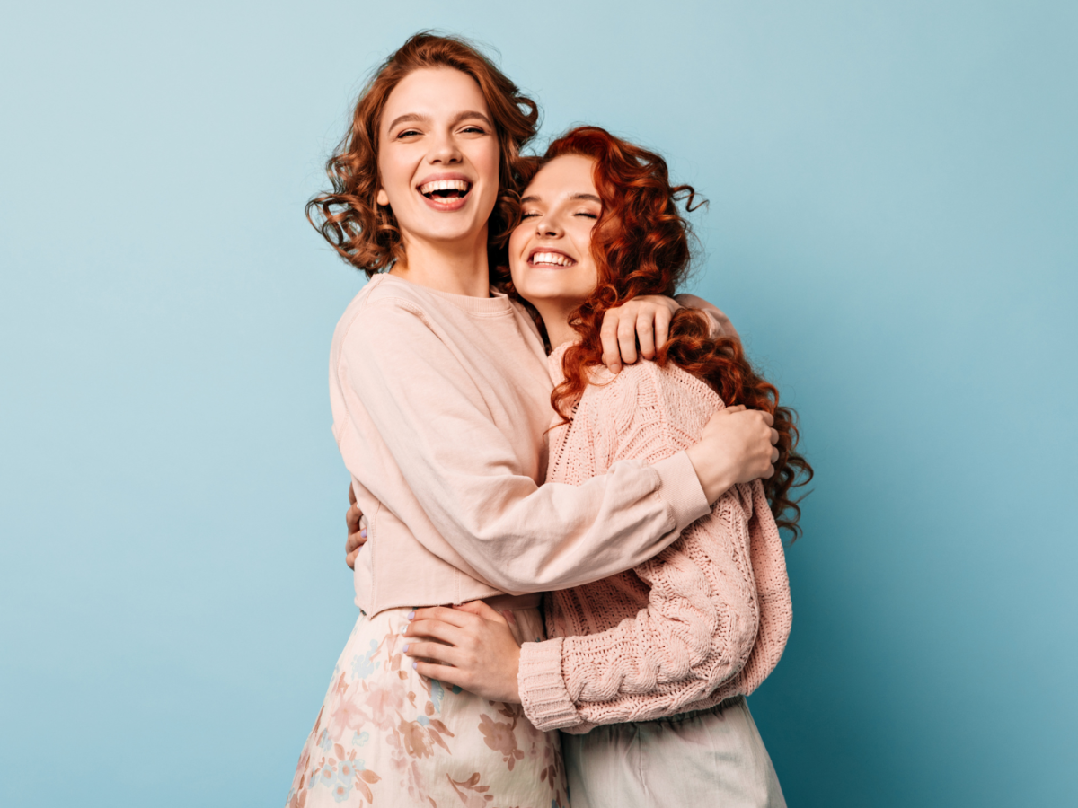 2 junge Frauen stehen vor einem hellblauen Hintergrund, freuen sich und nehmen sich in den Arm