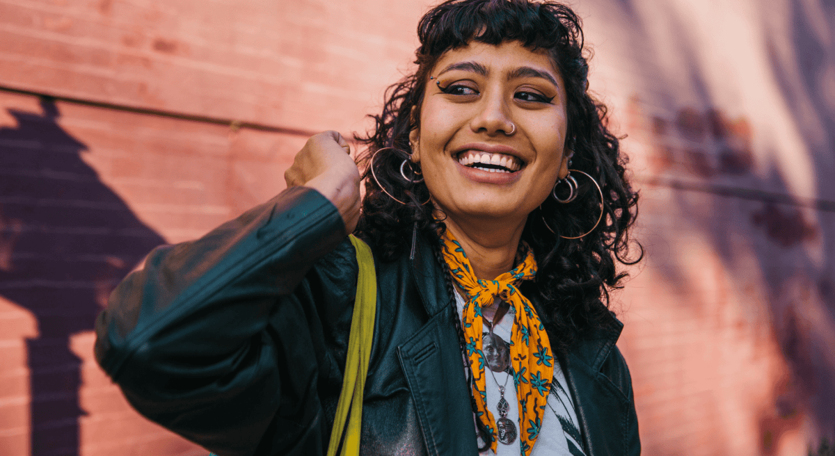 Frau mit dunklen Locken, Piercings und einem Halstuch steht vor einer pinken Wand und strahlt