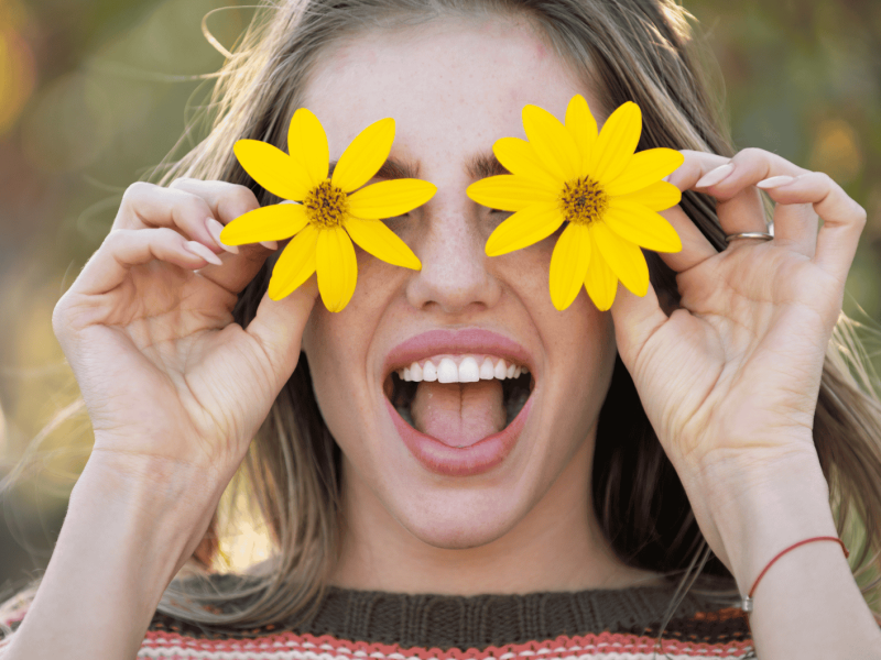 Frau lächelt mit offenem Mund und hält sich zwei gelbe Blüten vor die Augen