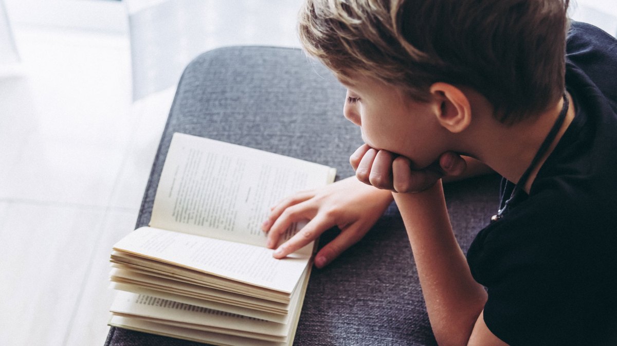 Junge, ca. 10 Jahre alt, liegt auf der Couch und liest ein Buch.