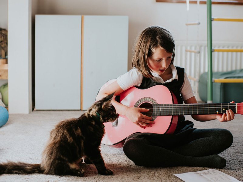 Mädchen sitzt auf dem Fußboden ihres Kinderzimmers und spielt auf einer rosafarbenen Gitarre. Vor ihr sitzt eine kleine Katze.