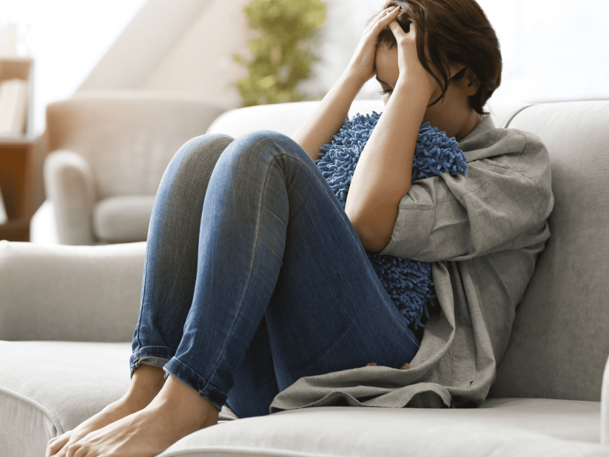 Frau sitzt mit angezogenen Beinen und Kopf in den H&auml;nden gest&uuml;tzt frustriert auf dem Sofa